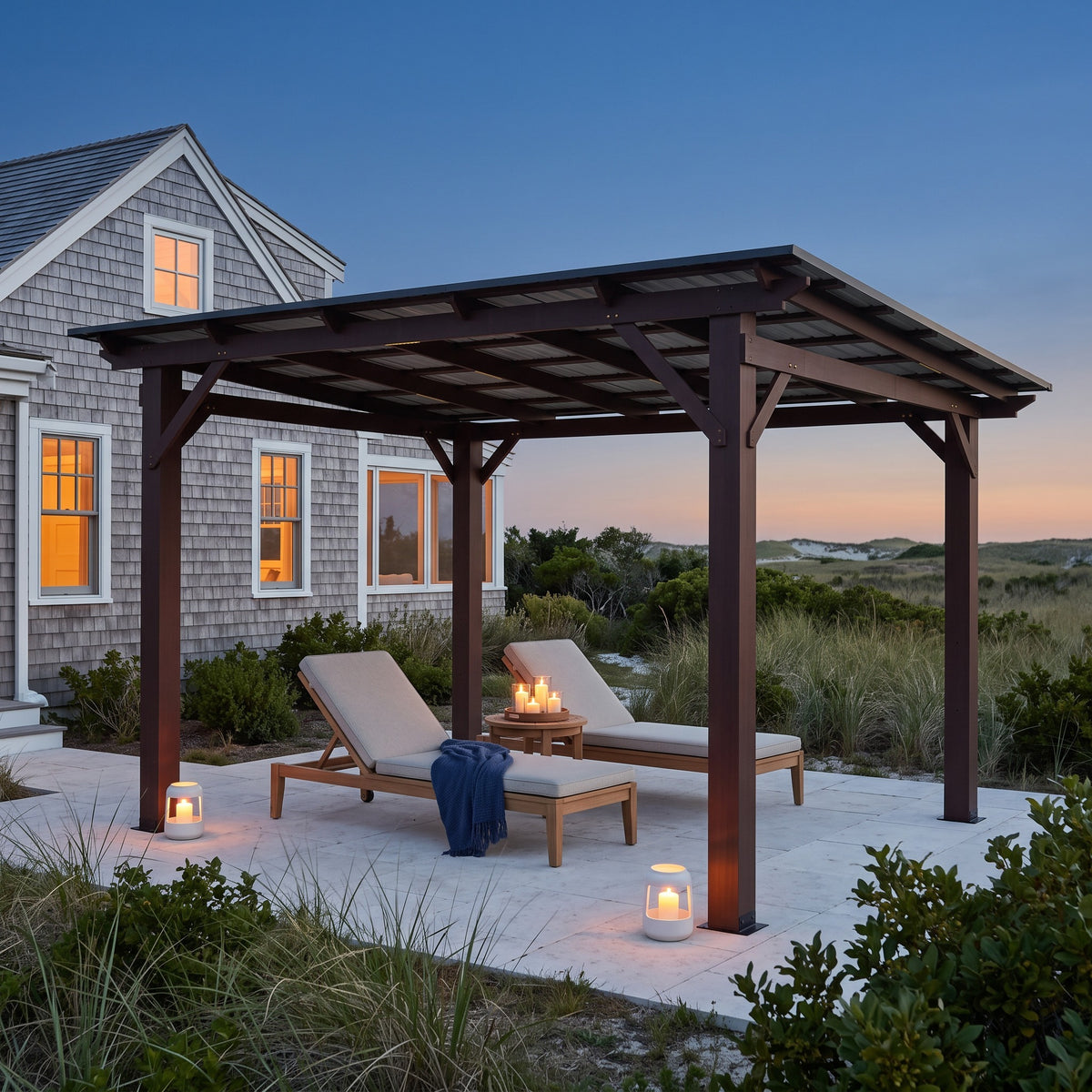Brown wood patio gazebo with a dark metal roof over two lounge chairs on a stone patio by a shingled house at dusk