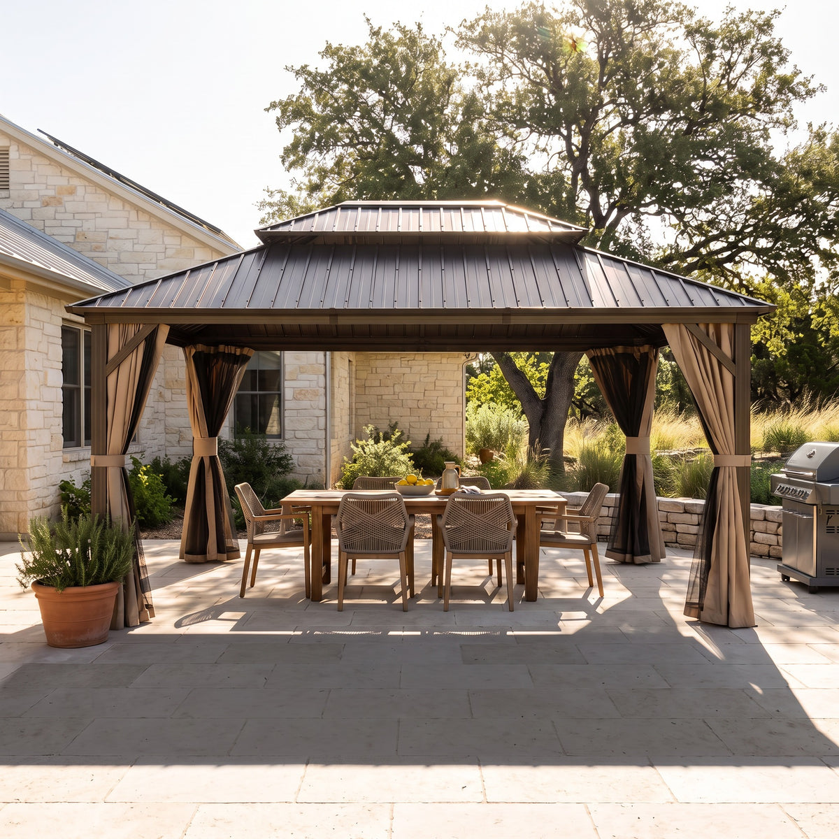 Brown aluminum 10x14 double roof hardtop gazebo with tan curtains over a wood dining table on a stone patio