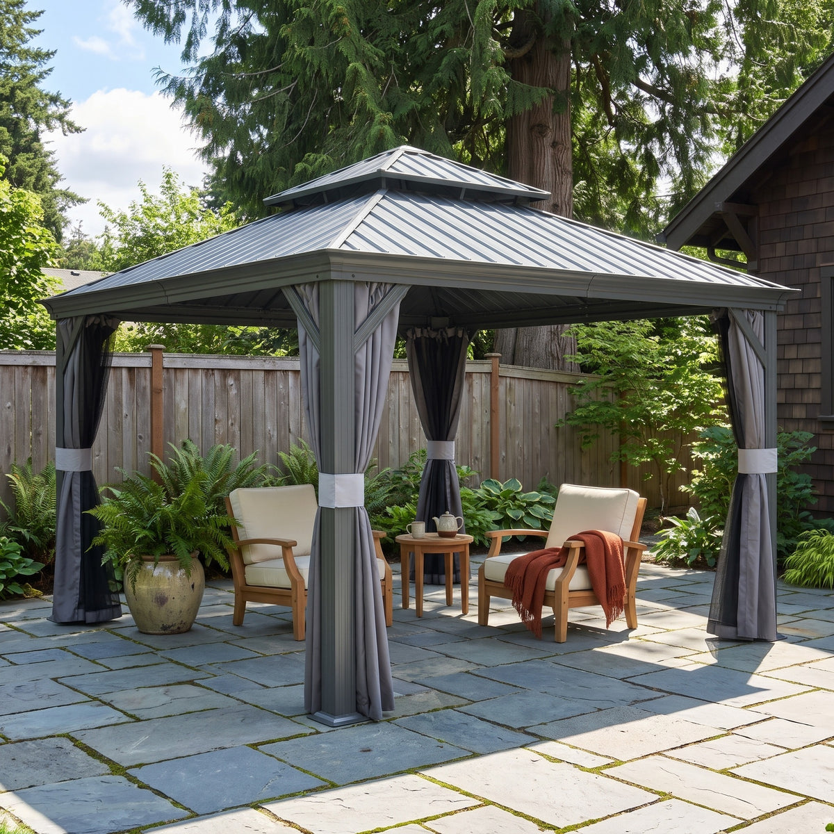 Gray aluminum 12x12 double roof hardtop gazebo with tied back curtains over two lounge chairs on a stone patio