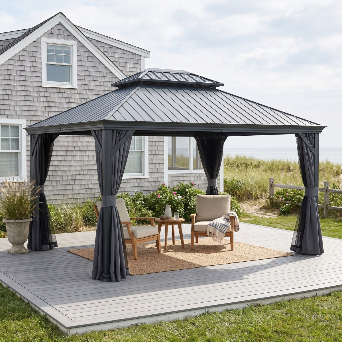 Dark gray 12x14 hardtop gazebo with tied back curtains over two lounge chairs on a gray deck beside a shingled house