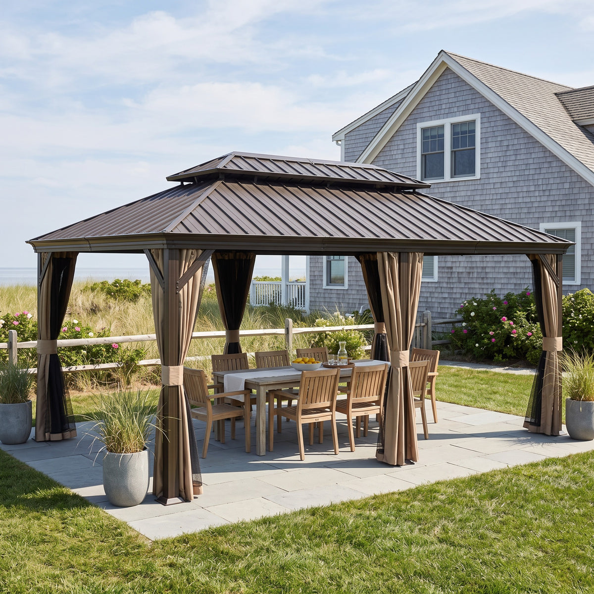 Brown 12x18 hardtop gazebo with tan curtains and netting over a wood dining set on a stone patio by a coastal house
