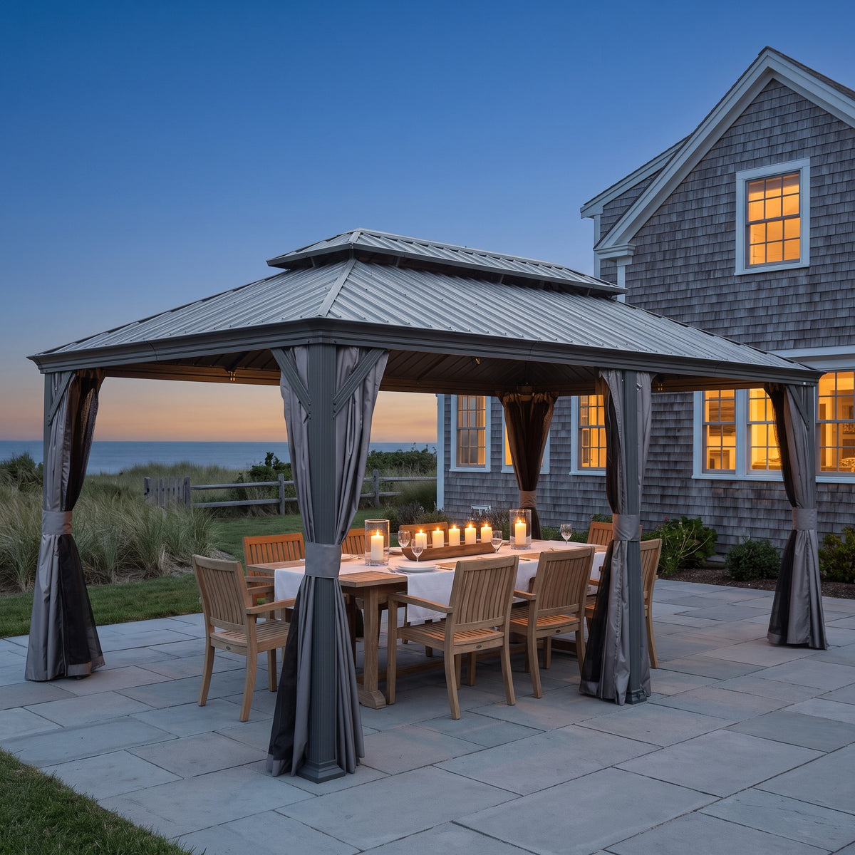 Gray 12x18 hardtop gazebo with tied-back curtains over a candlelit wood dining table on a stone patio at dusk