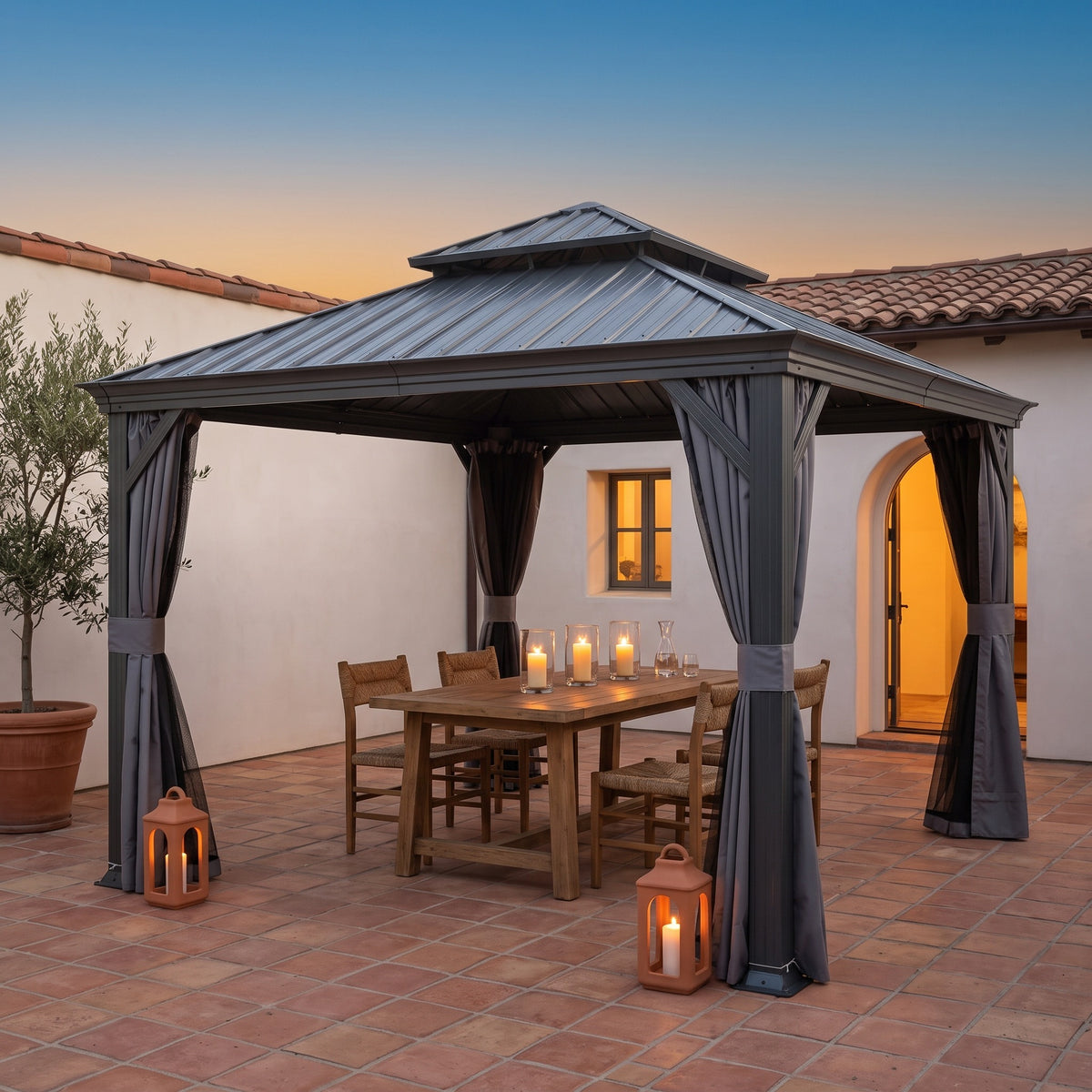 Gray steel double roof hardtop gazebo with tied back curtains over a candlelit dining table in a tiled courtyard at dusk