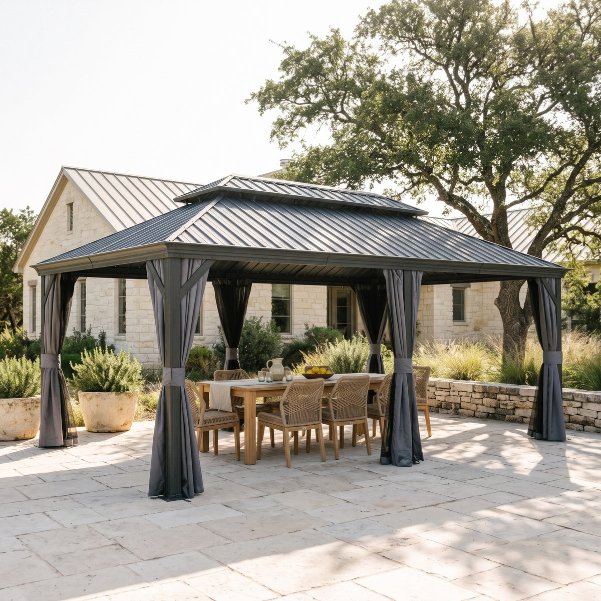 Gray 14x20 hardtop gazebo with tied back curtains over a teak dining table on a stone patio in daylight