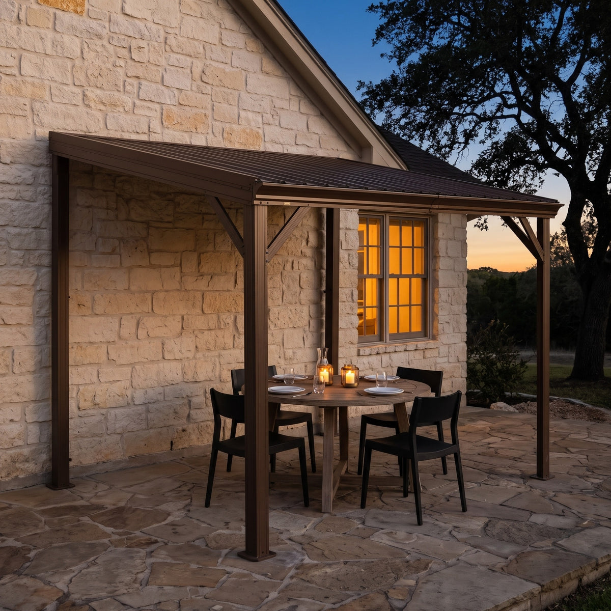 Brown metal lean-to hardtop gazebo against a stone house over a round dining table lit by candle lanterns at dusk