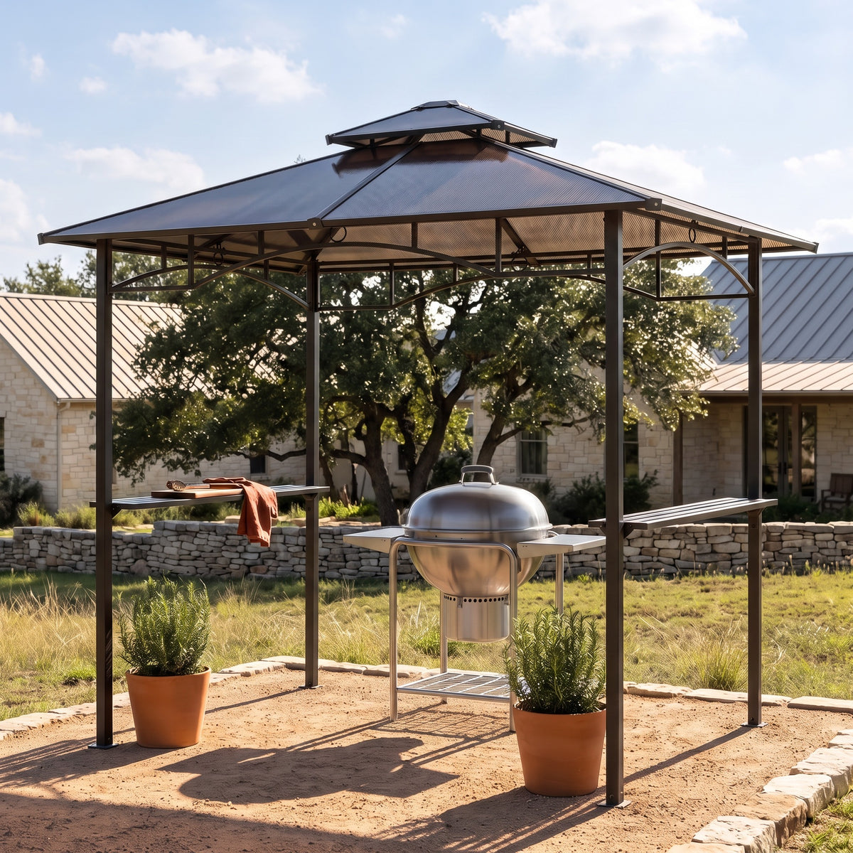 Brown metal grill gazebo with double roof and two side shelves over a kettle grill on a sandy patio by a stone house