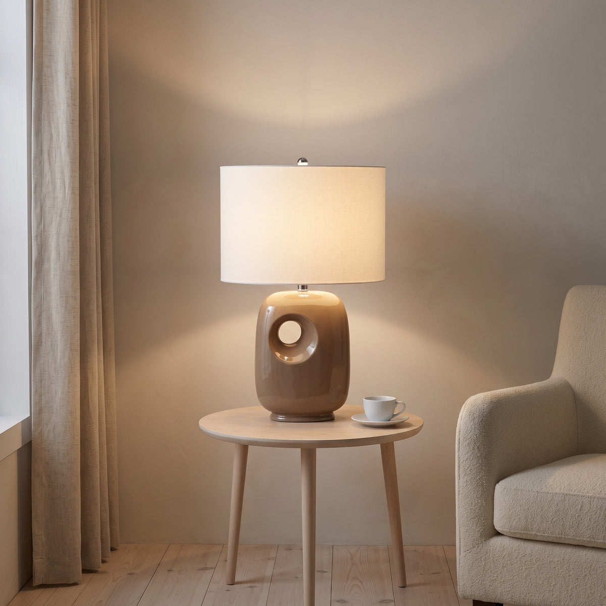 Brown ceramic table lamp with cut-out base and white linen-look drum shade, lit, on a round oak side table