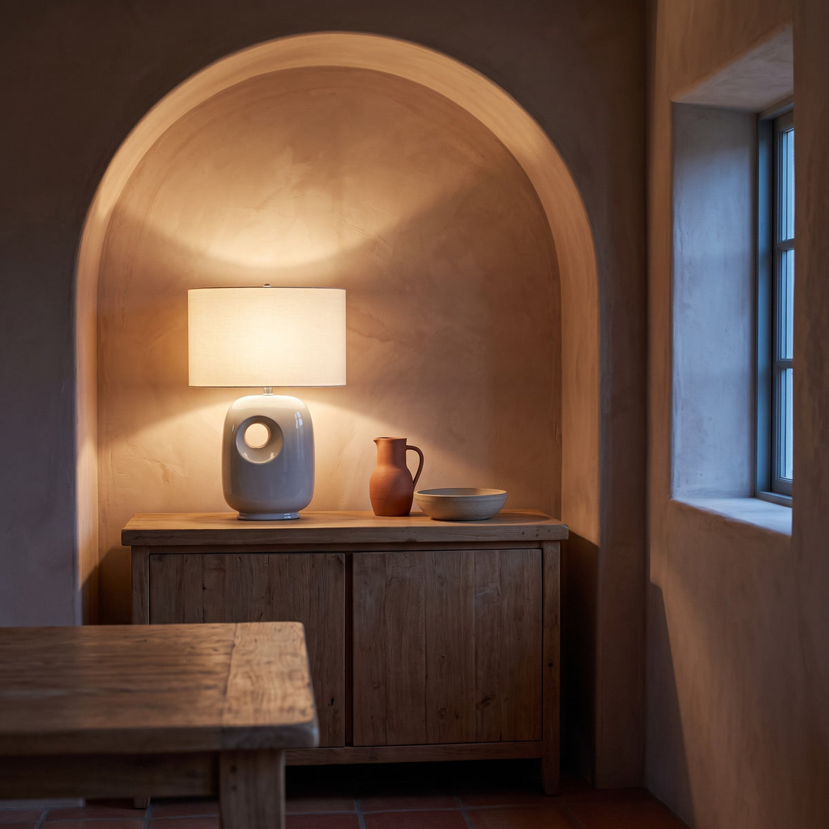 Grey ceramic table lamp with cut-out base and lit ivory shade on a wood cabinet in an arched plaster alcove