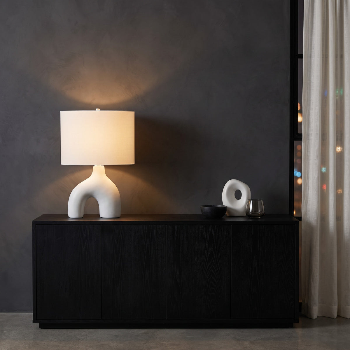 White ceramic table lamp with arched base and ivory fabric drum shade, lit, on a black wood console