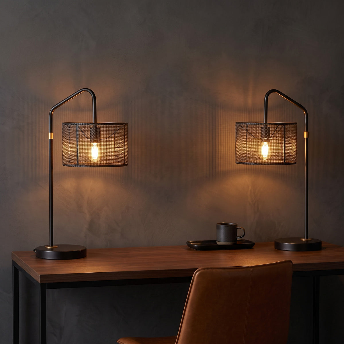 Pair of bronze table lamps with black metal mesh drum shades, lit, on a walnut desk against a dark wall