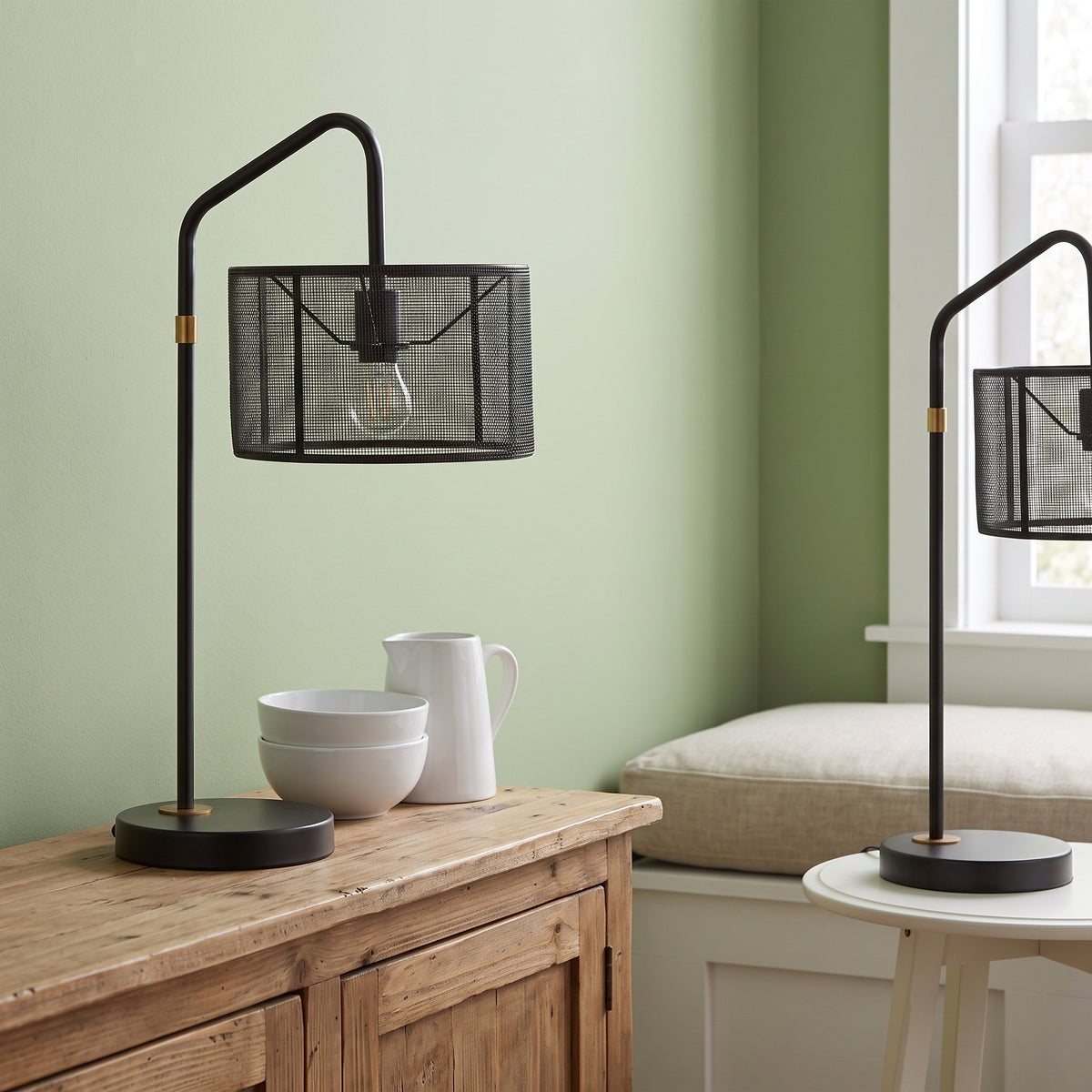 Two black metal mesh shade table lamps with gold accents on a wood cabinet and white side table by a window