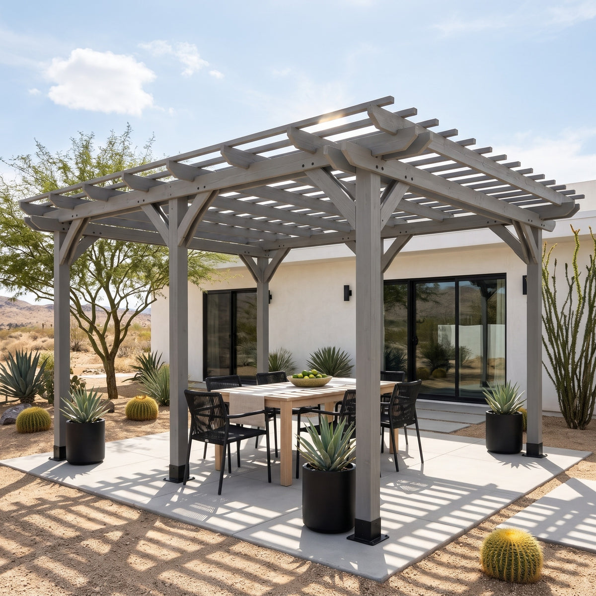 Gray wood 12x16 pergola over a dining table on a concrete patio beside a white desert home in sunlight