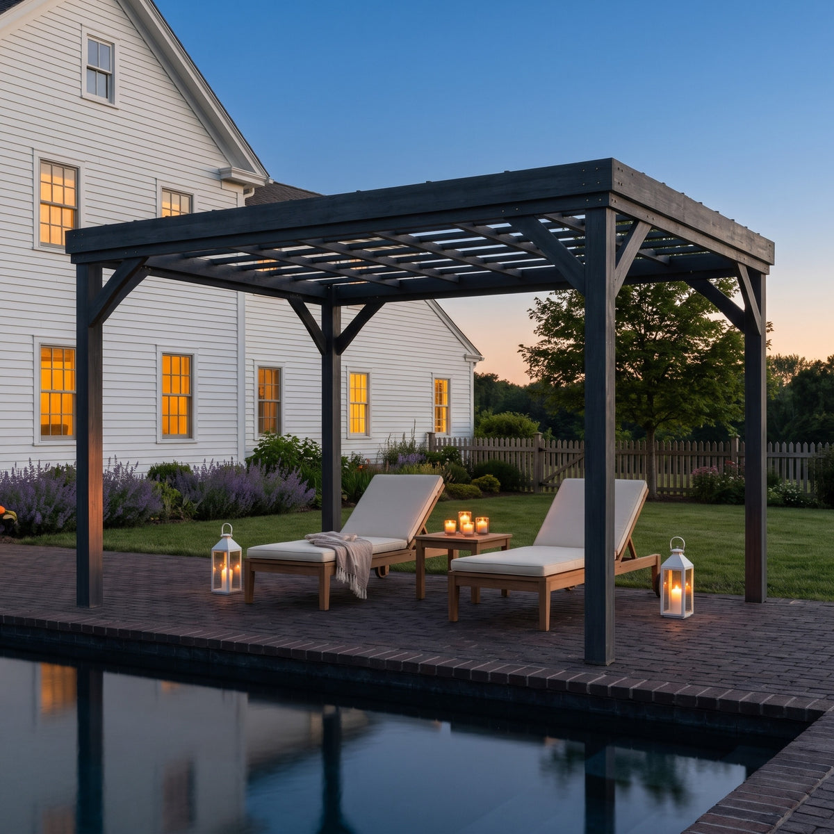 Gray wood pergola with open grid roof over two lounge chairs by a pool at dusk, lit by lanterns and candles