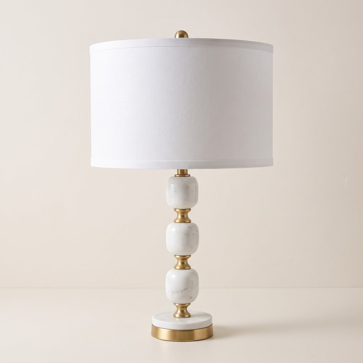 White marble table lamp with brass accents and white drum shade on an ivory backdrop