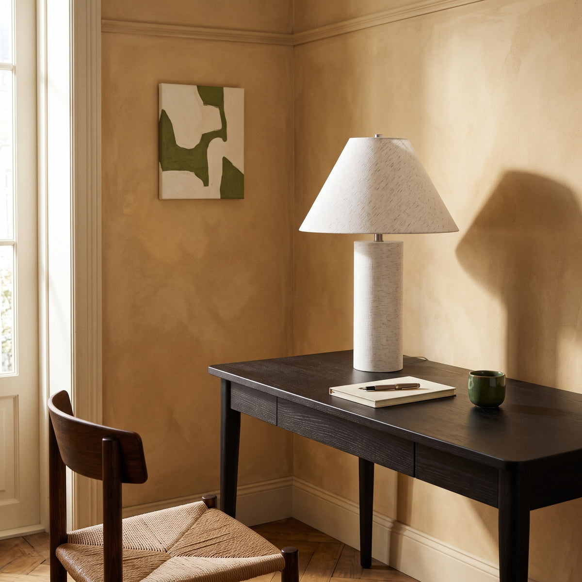White table lamp with linen shade on a dark wood desk in a sunlit study corner