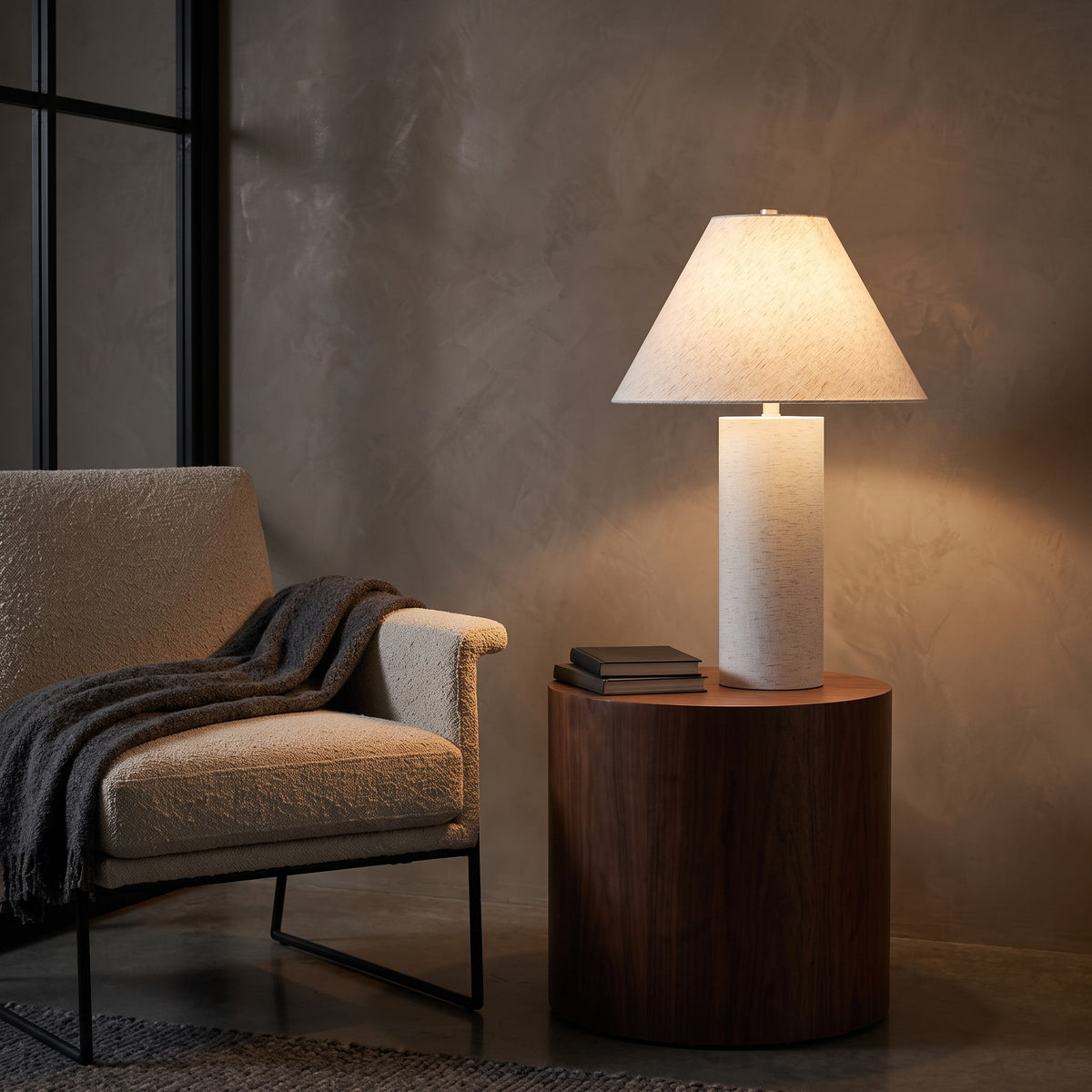 White table lamp with linen shade glowing on a round wood side table beside a lounge chair