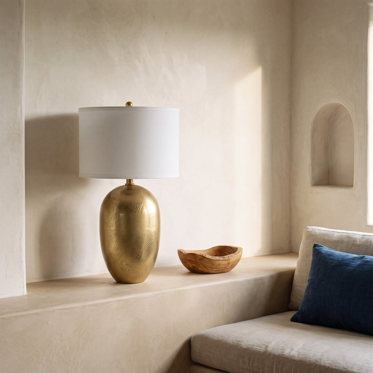 Gold table lamp with crosshatch etched body and white drum shade on a plaster ledge beside a linen sofa