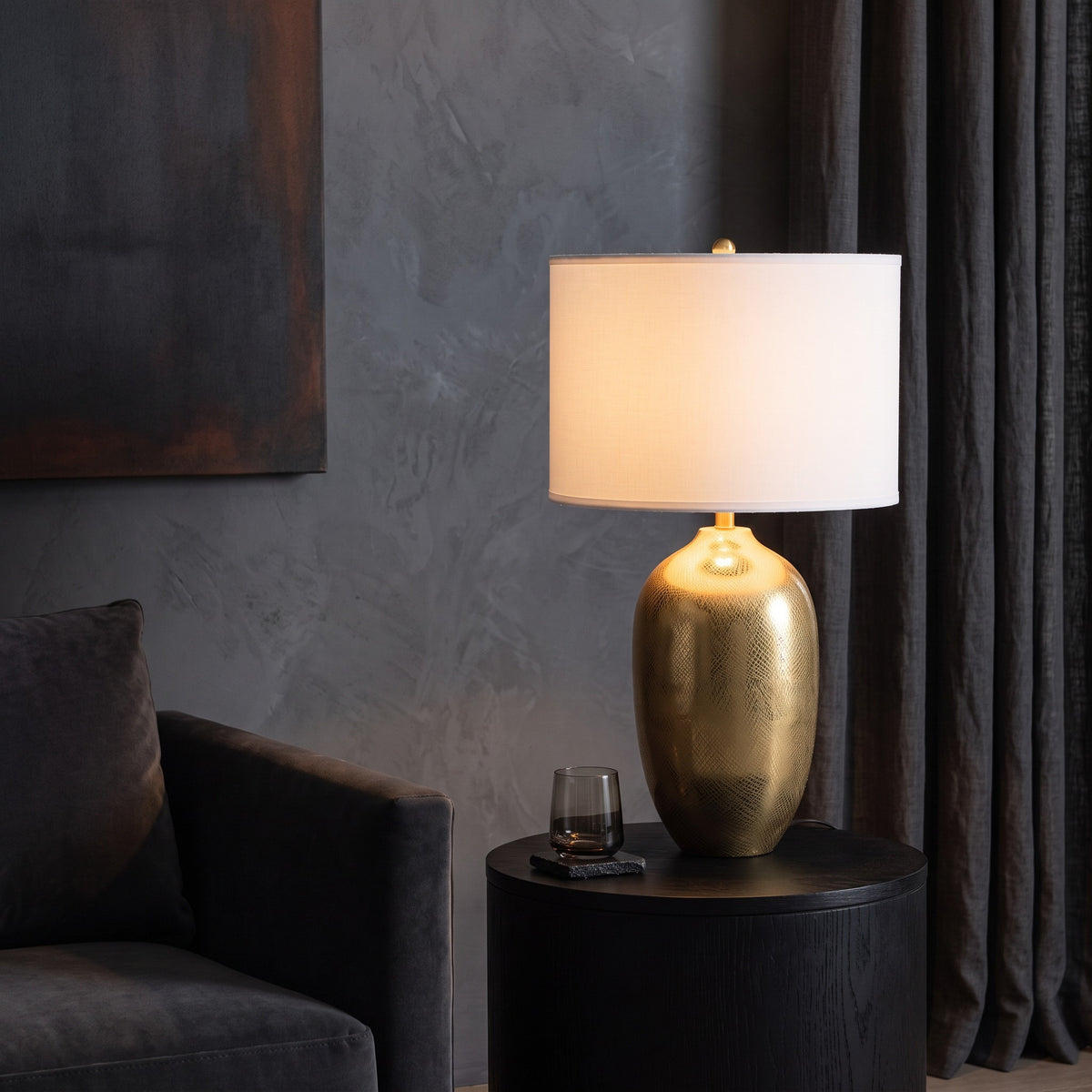 Gold etched urn table lamp with white drum shade lit on a round black side table beside a gray velvet armchair