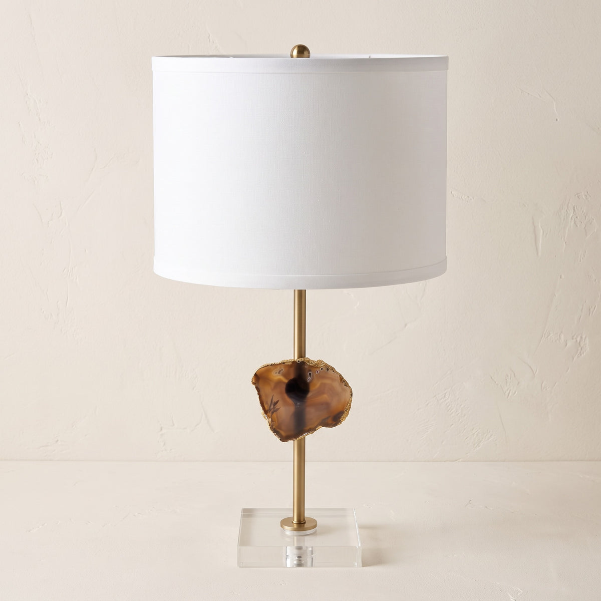 Table lamp with white drum shade, brass stem, gold-edged agate slice and clear acrylic base on an ivory plaster backdrop