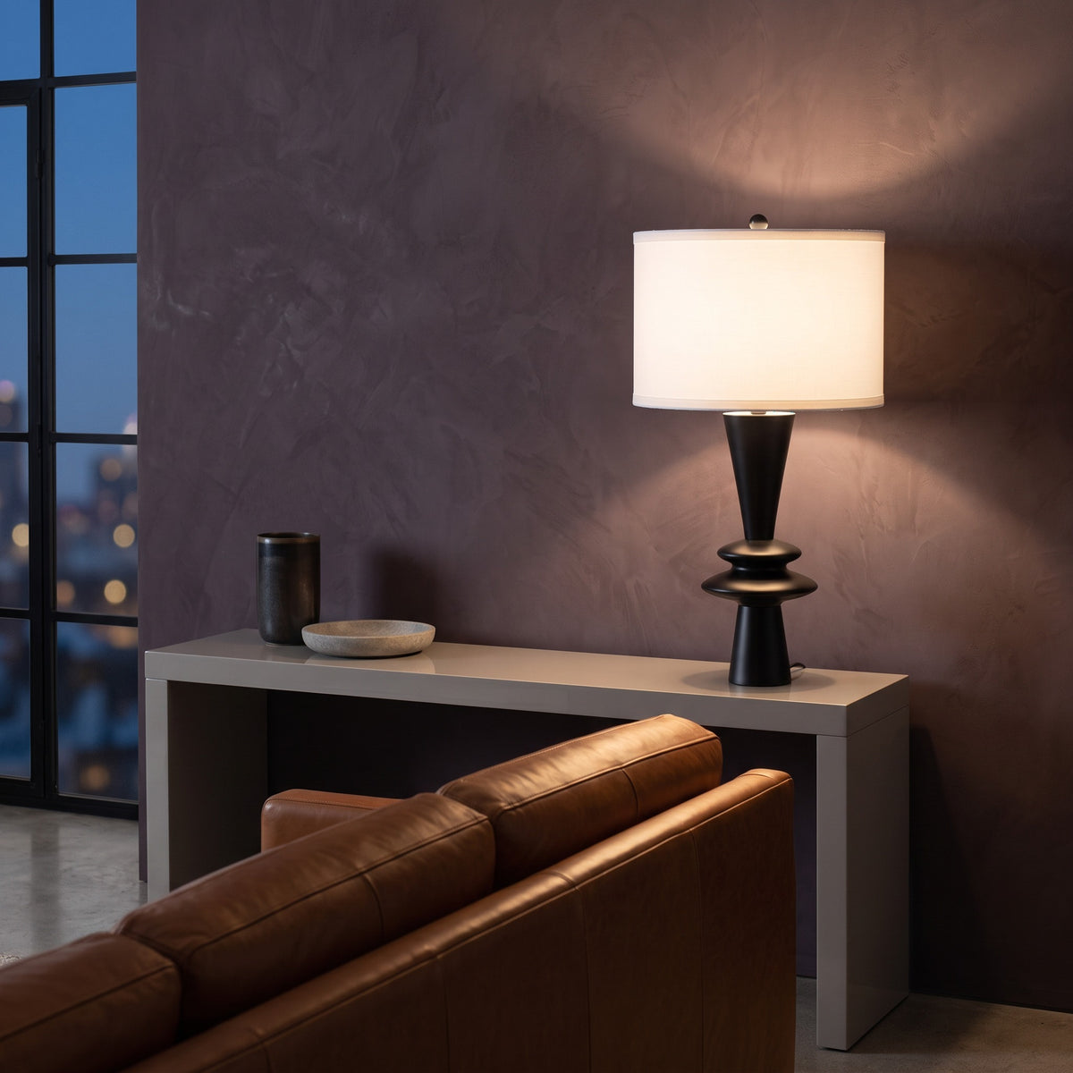 Black table lamp with stacked disc base and white drum shade lit on a gray console at night