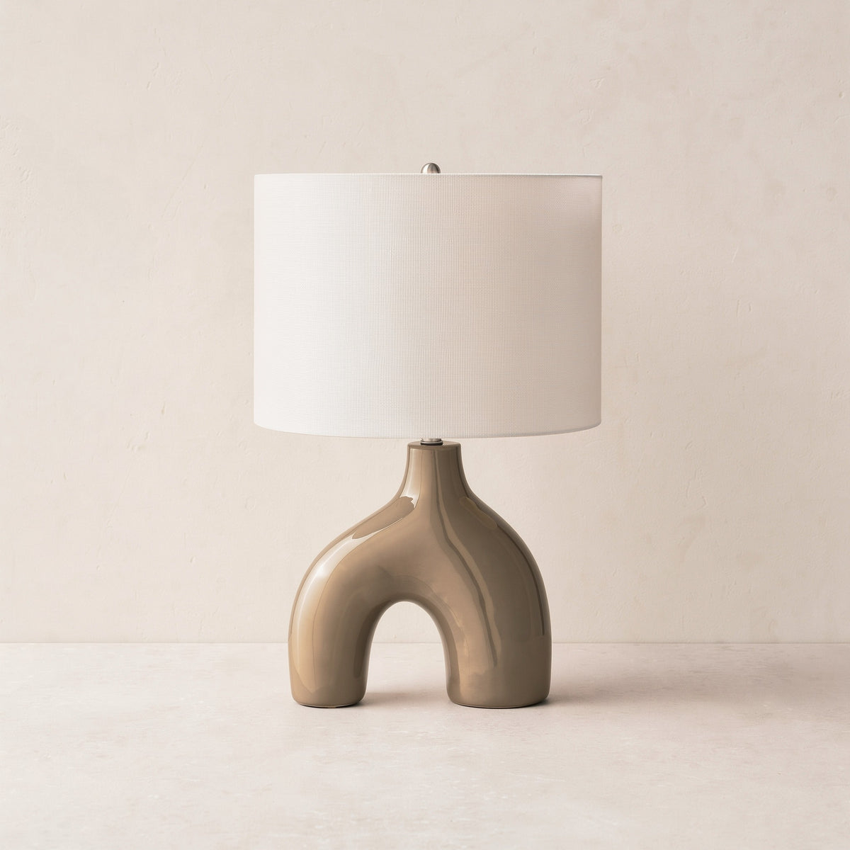 Brown glossy ceramic arch table lamp with white drum fabric shade on an ivory plaster backdrop
