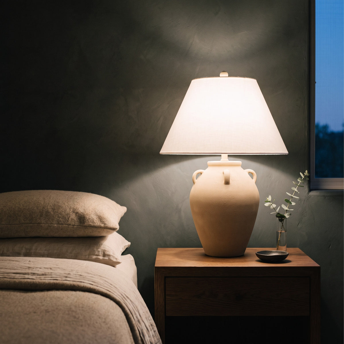 Beige ceramic jar table lamp with white linen-look shade, lit on a walnut nightstand beside a bed