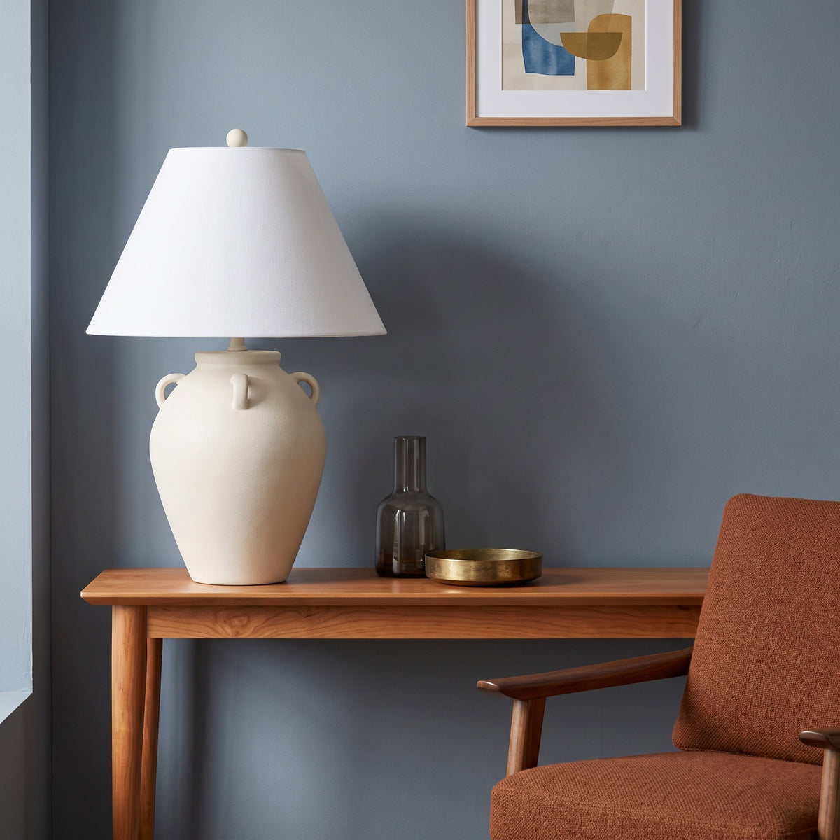 Cream ceramic jar table lamp with loop handles and white linen-look shade on a cherry console against a blue wall