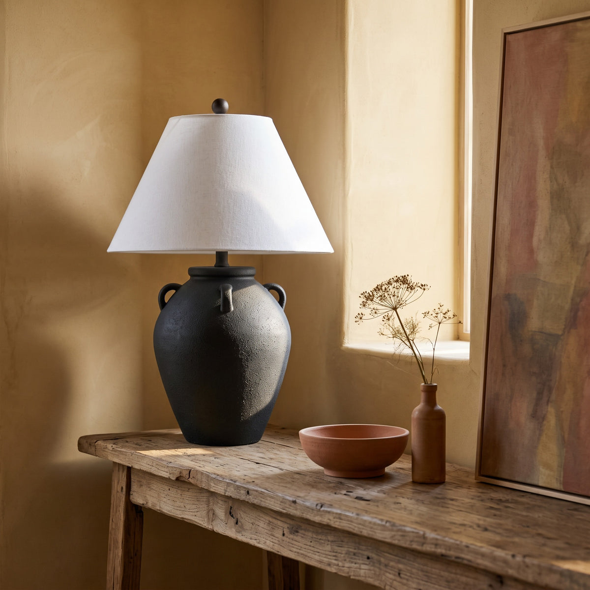 Black textured ceramic jar table lamp with white linen shade on a rustic wood console by a sunlit window