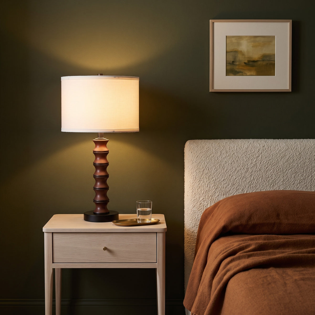 Cherry spool table lamp with white linen-look drum shade lit on an oak nightstand beside a bed