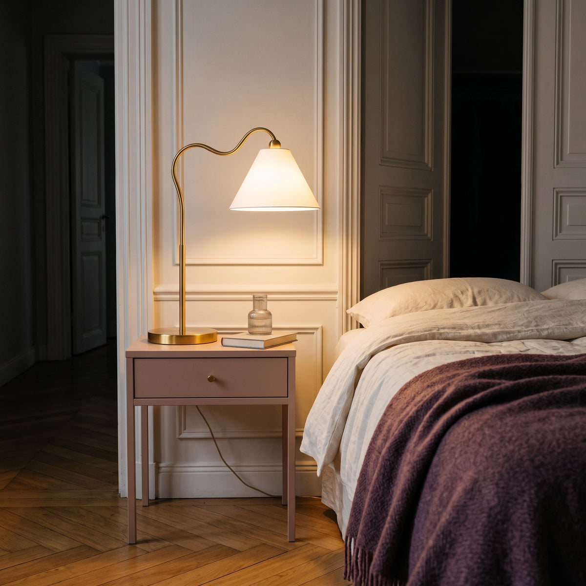 Gold table lamp with curved arm and white cone shade lit on a blush nightstand beside a linen bed