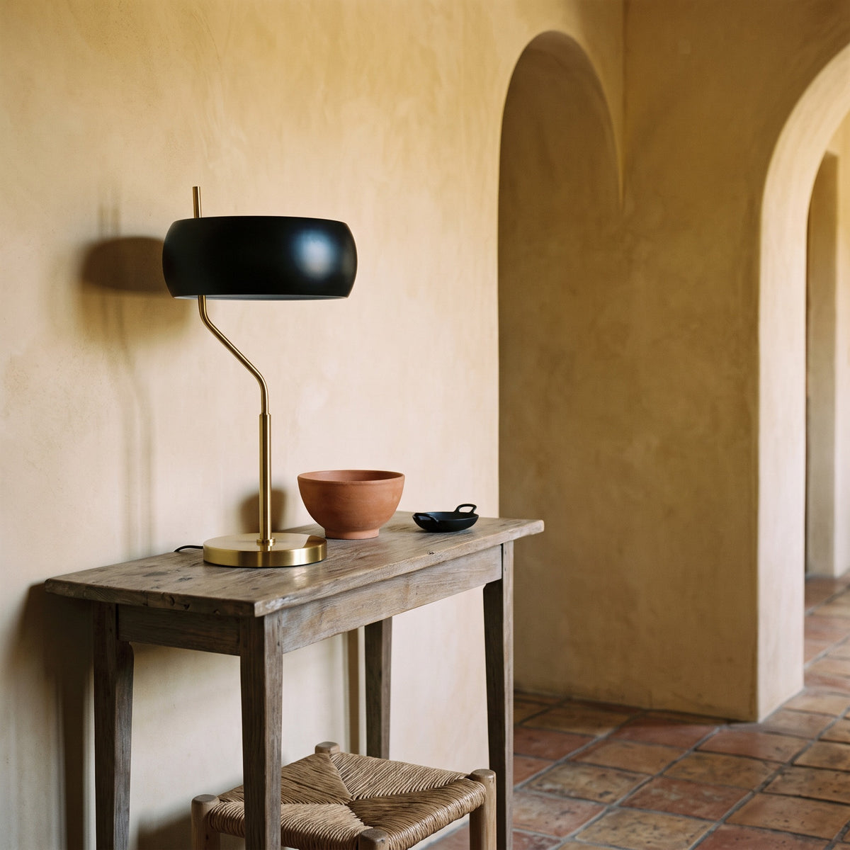 Black and gold table lamp with drum shade on a rustic wood console beside a terracotta bowl in an arched plaster hall