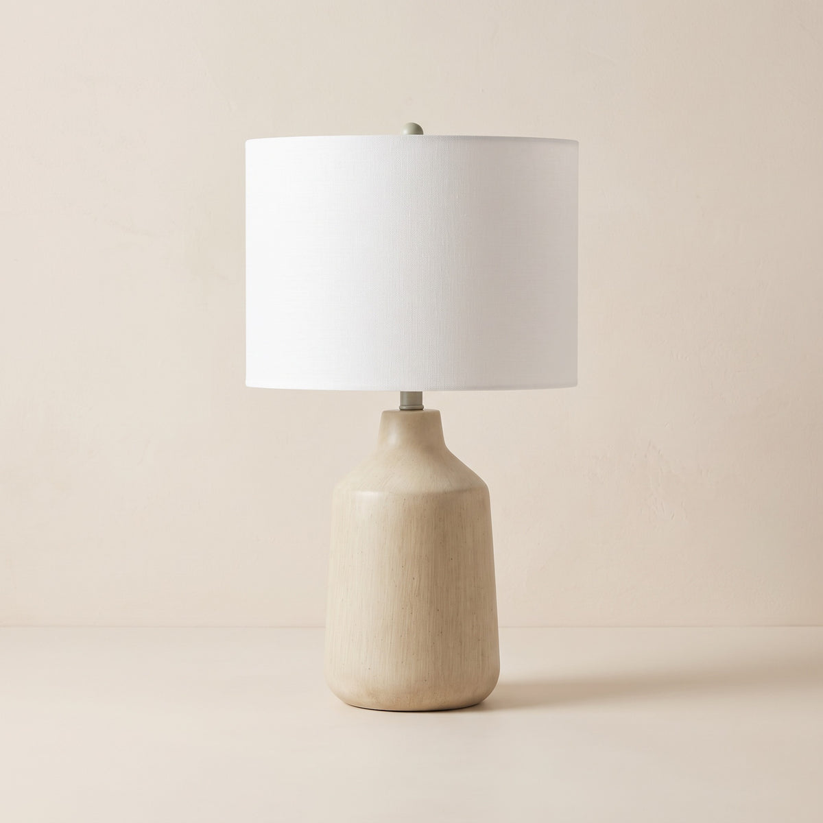 Beige concrete table lamp with white linen drum shade and sage finial on an ivory plaster backdrop