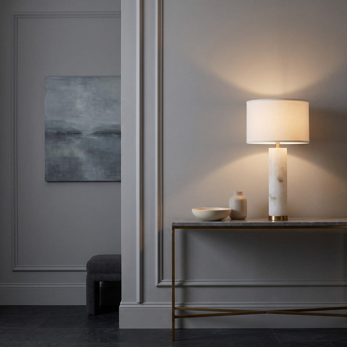 White alabaster table lamp with gold base and white drum shade, lit on a marble top console in a paneled hallway