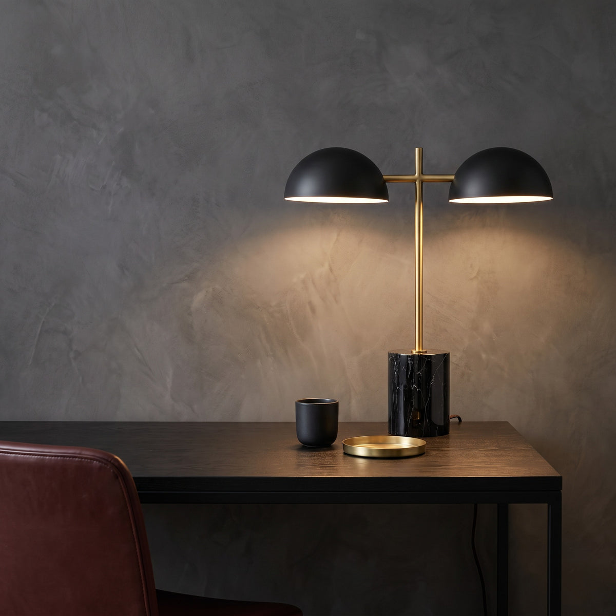 Black marble-look table lamp with two black dome shades and brass finish stem, lit on a dark wood desk by a plaster wall