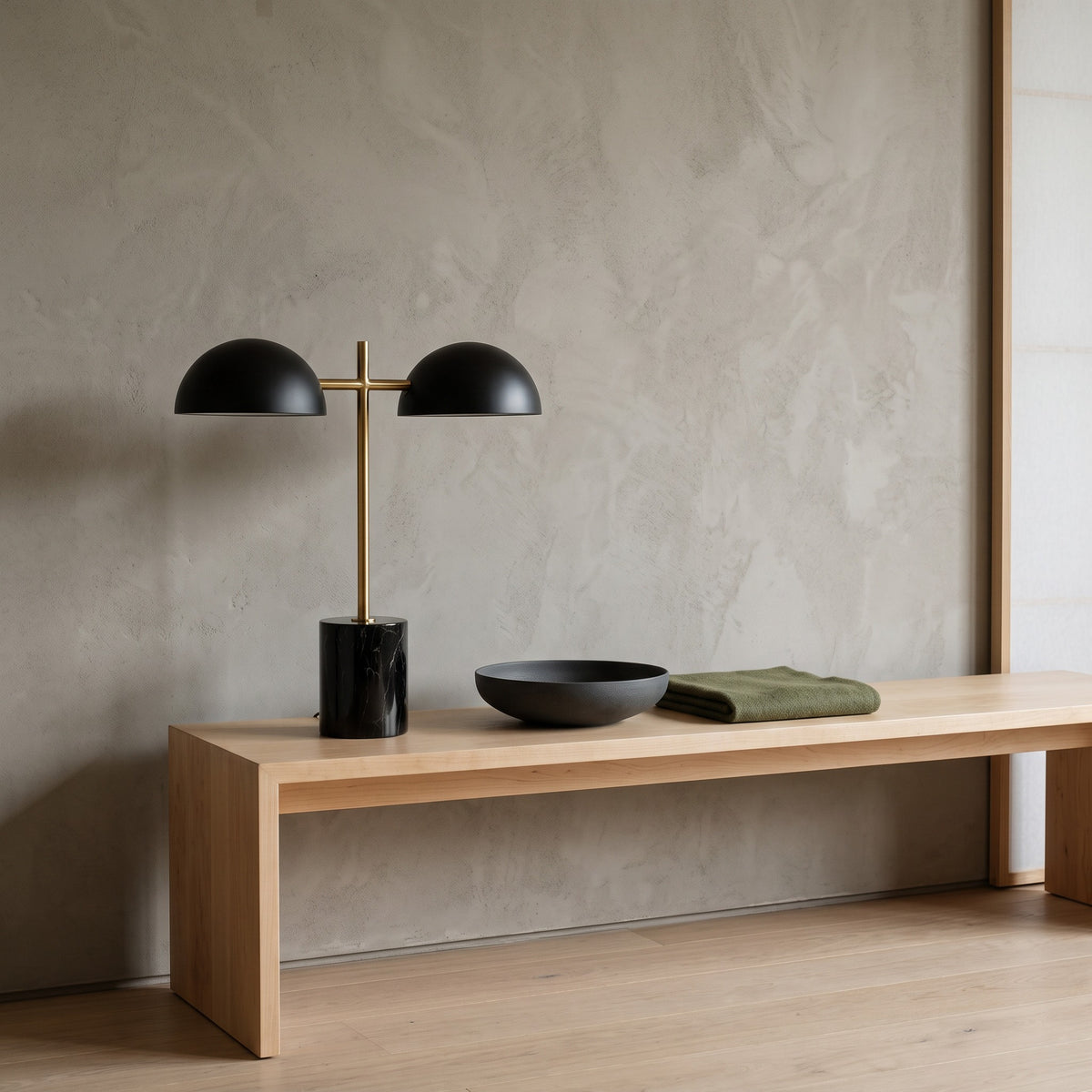 Black marble-look table lamp with two black dome shades and brass finish stem on a light oak bench by a plaster wall