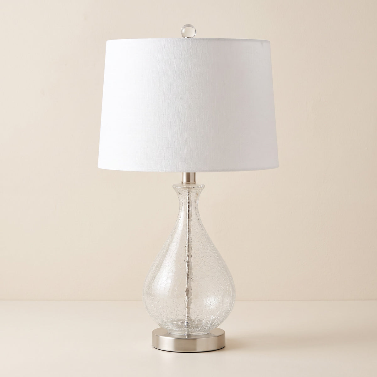 Clear crackle glass table lamp with white linen drum shade and nickel base on an ivory backdrop