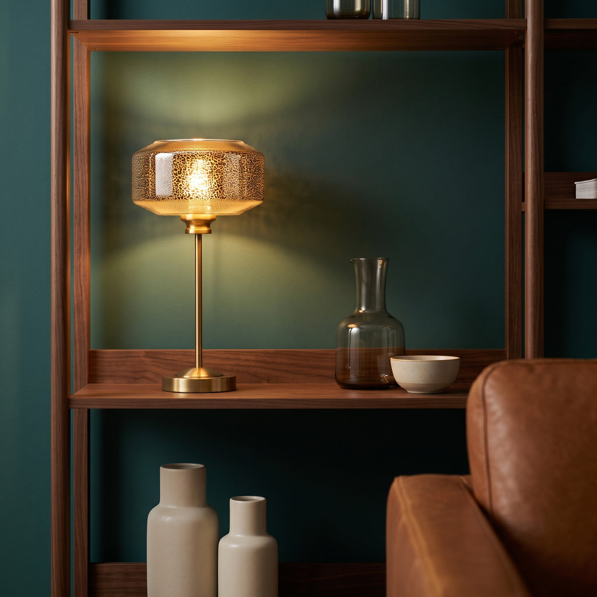Gold table lamp with mercury glass shade lit on a walnut bookshelf beside a glass carafe and ceramic bowl
