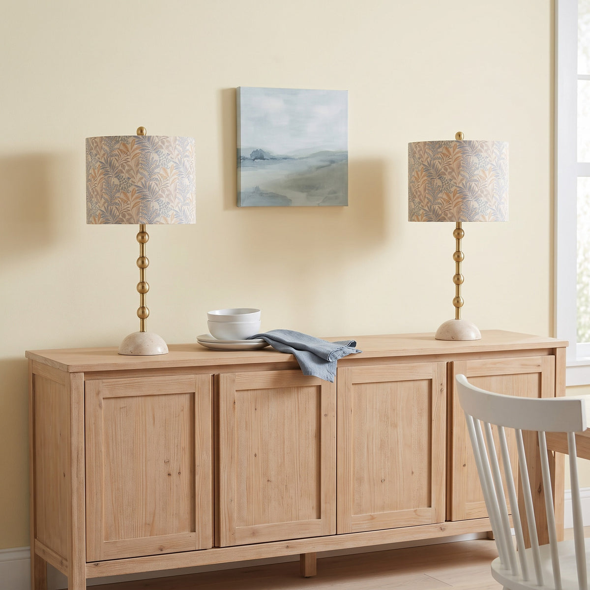 Pair of gold table lamps with travertine bases and leaf print drum shades on a light wood sideboard