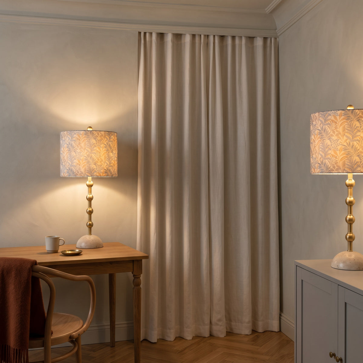 Pair of gold table lamps with travertine bases and leaf print drum shades, lit on a desk and white cabinet