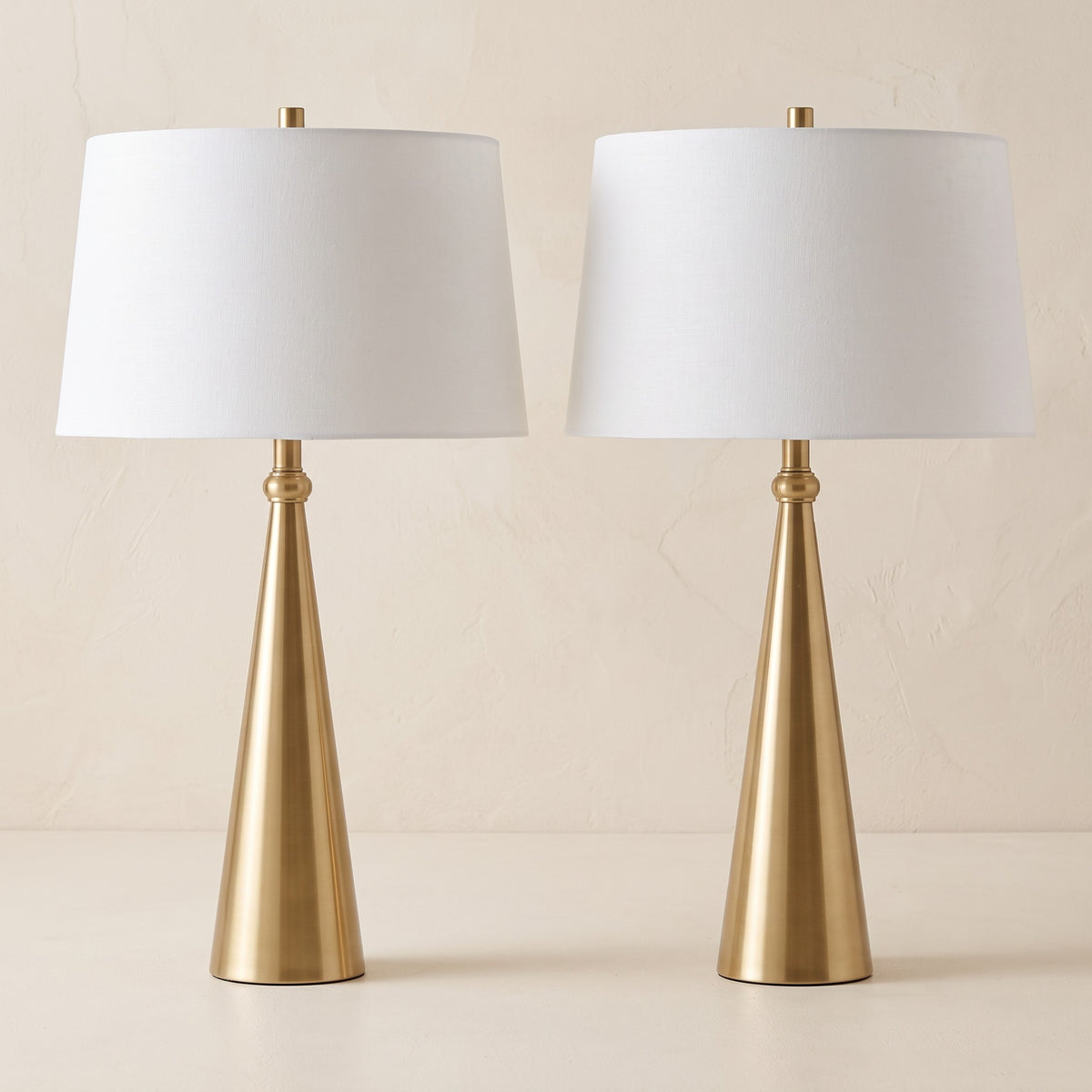 Pair of gold cone table lamps with white linen tapered shades against an ivory plaster wall