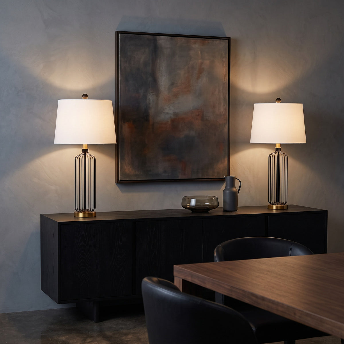 Two black wire cage table lamps with brass bases and white shades lit on a black sideboard in a dining room