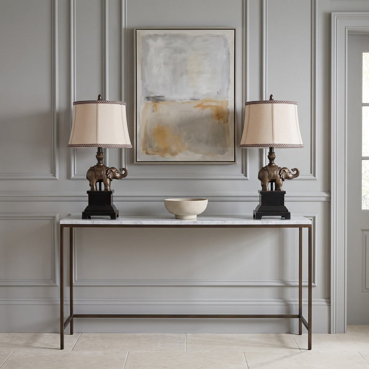 Pair of bronze elephant table lamps with natural linen shades on a marble console in a grey panelled hallway