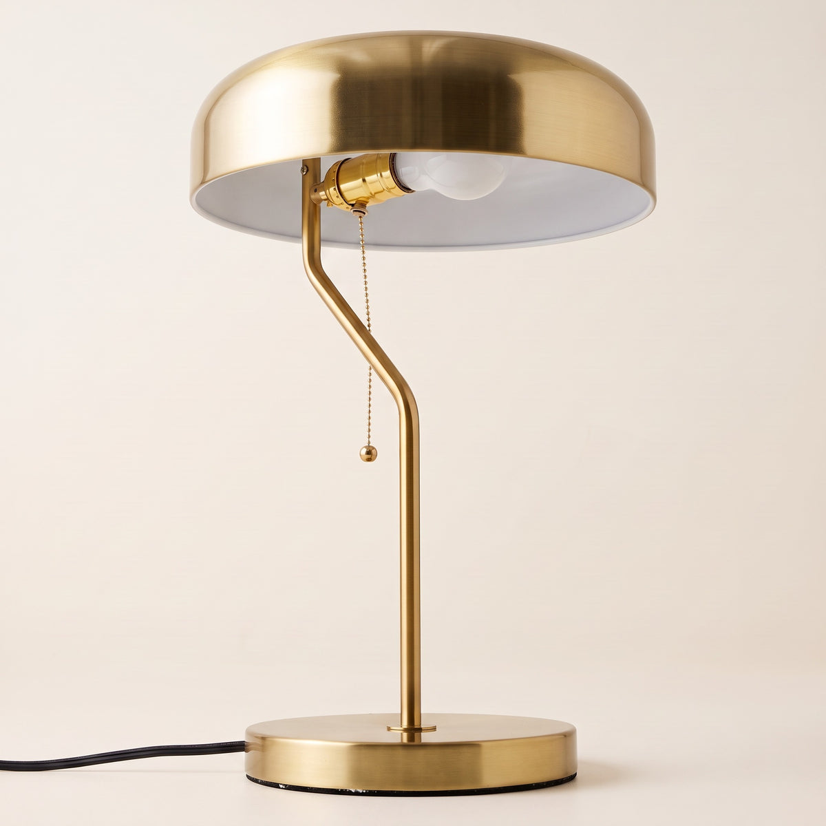 Brushed gold table lamp with dome metal shade, offset curved stem and pull chain on an ivory backdrop