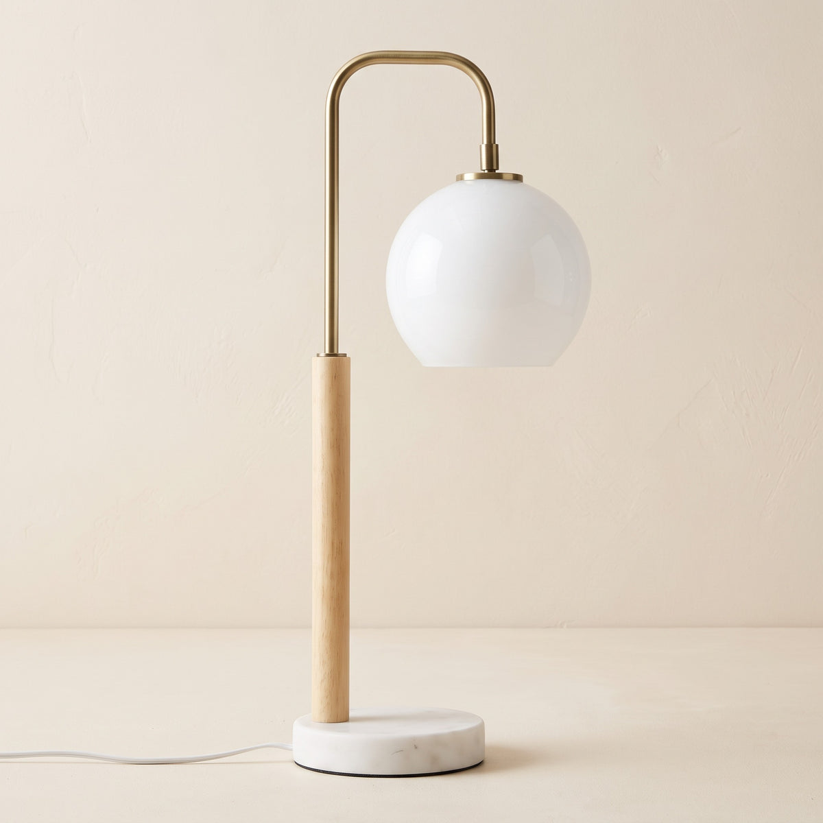 White globe table lamp with brass finish arm, light wood stem and white marble base on an ivory backdrop