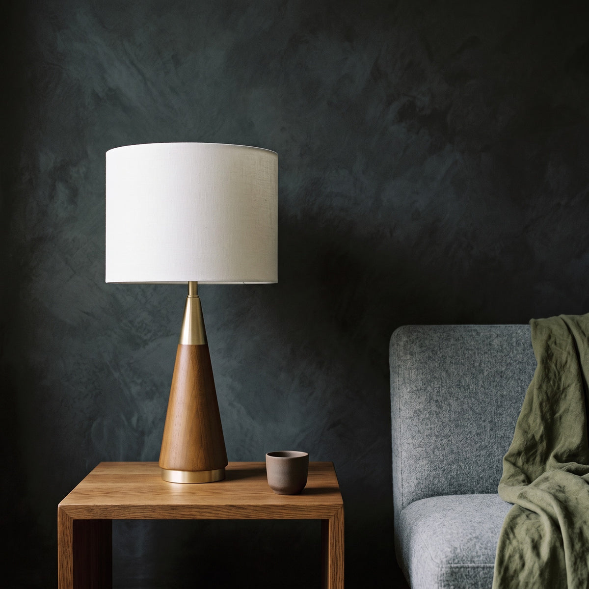 Wood and brass cone table lamp with white linen-look drum shade on an oak side table beside a gray armchair