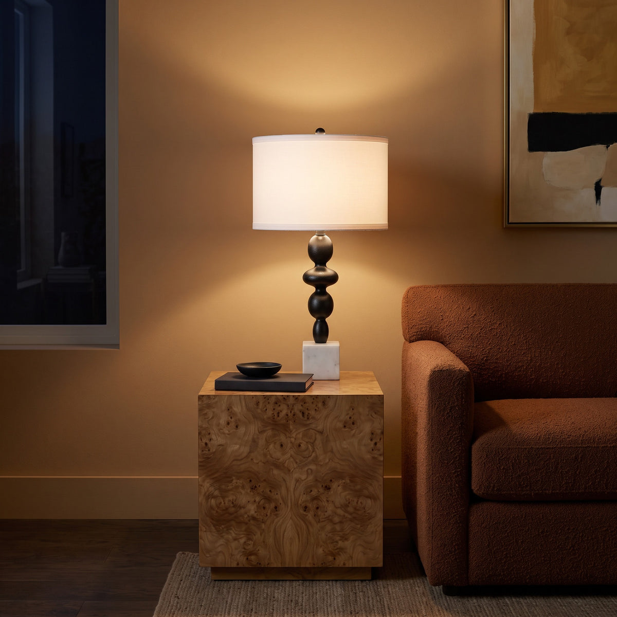 Black and white marble table lamp, lit, with white drum shade on a burl wood end table beside a rust boucle sofa