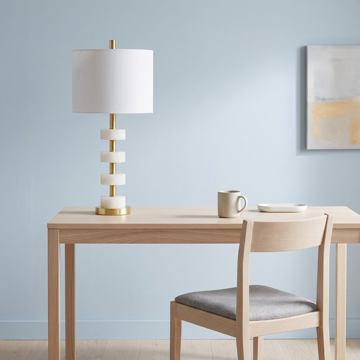 White marble and brass stacked disc table lamp with white drum shade on a light oak desk against a blue wall