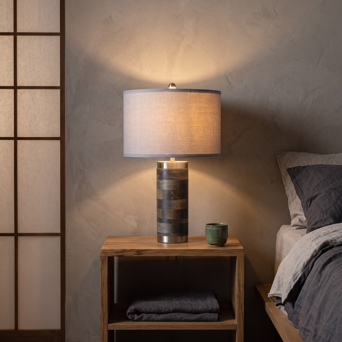 Gray drum shade table lamp with banded wood base, lit, on an oak nightstand beside a linen bed